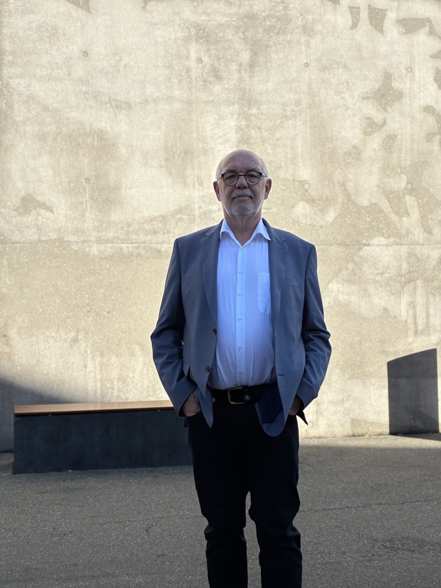 Detlef Durdel steht in Sakko und weißem Hemd vor einer hellen Betonwand, die Hände in den Hosentaschen