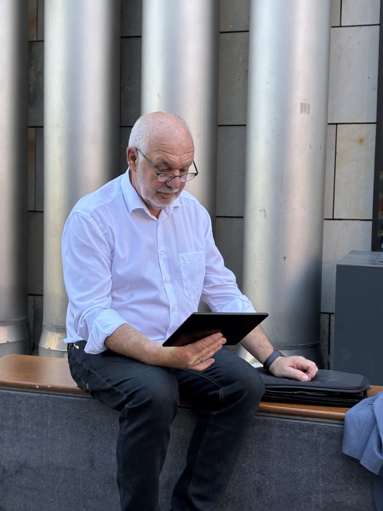 Detlef Durdel sitzt auf einer Bank vor glänzenden Metallrohren und liest auf einem Tablet